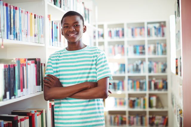 A schoolboy is standing in a library with his arms crossed, smiling confidently. He is surrounded by bookshelves filled with books, indicating an academic environment. This image can be used for educational materials, school brochures, library promotions, or articles about student life and learning.