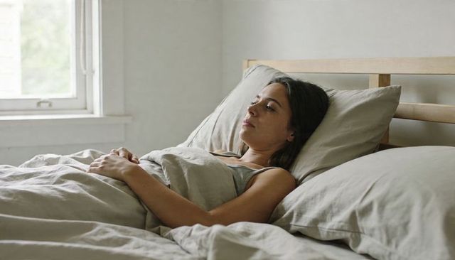 Young woman resting in neutral linen bedding, contemplative morning, natural light, calm bedroom