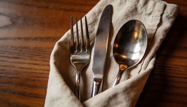 Stainless steel fork knife spoon nestling in folded beige linen napkin on wooden table