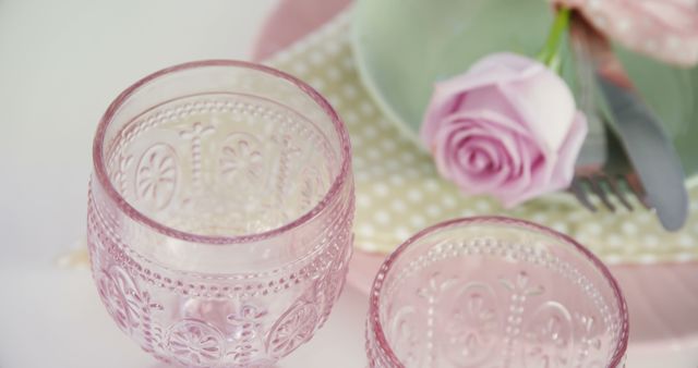 Elegant pink glassware is showcased near a delicate floral arrangement, with copy space. The soft hues and intricate patterns suggest a setting prepared for a refined event or celebration.