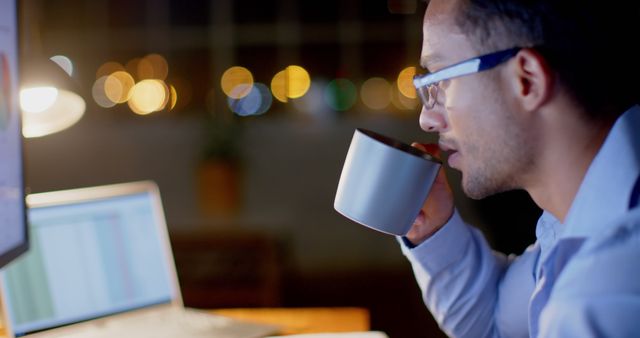 Person working late night, focused on laptop screen while drinking coffee. Bokeh background adds a soft, warm light ambiance implying nighttime hours. Ideal for use in articles about productivity, late-night working habits, technology integration in work, or promotional content for coffee and work-life products.