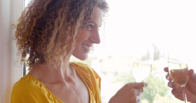 Woman with curly hair in yellow dress holding wine glass, smiling cheerfully during daylight. Perfect for depicting joyful celebrations, social events, casual parties, or lifestyle content.