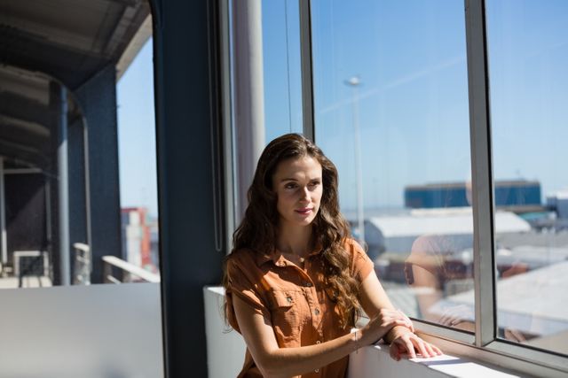 Businesswoman standing by a large window in a modern office, looking thoughtful. Ideal for use in corporate websites, business blogs, articles on workplace environments, or promotional materials for office spaces. The natural light and reflective surface add a contemplative mood, suitable for themes of professional reflection, decision-making, and business strategy.