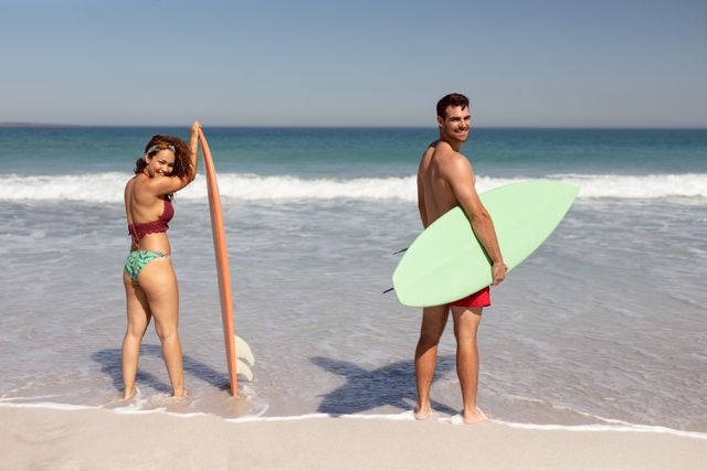 Rear view of happy young diverse couple with surfboard looking at camera on beach in the sunshine