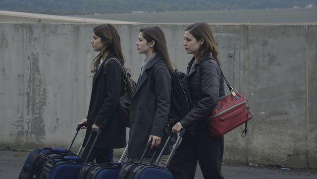 Three women walking pulling navy suitcases along concrete wall carrying backpacks red bag