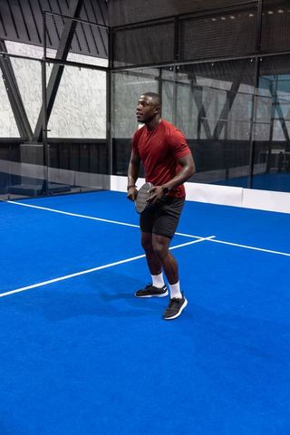 Athletic african american man playing paddle tennis on blue indoor court wearing red shirt