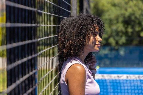 Curly-haired woman leaning on fence beside bright blue tennis court checking smartwatch