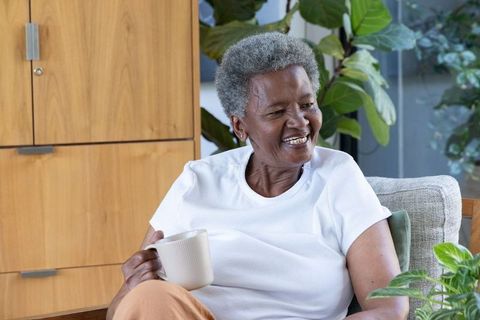 Senior african american woman smiling while holding mug in bright living room with plants