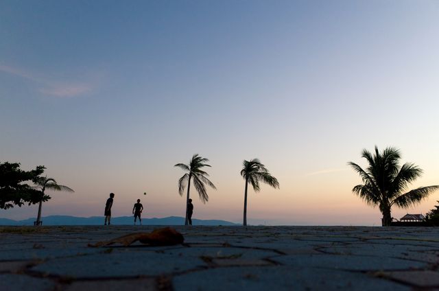 Silhouettes of children playing soccer at sunset on a tropical beach with palm trees in the background. This serene scene evokes childhood joy and simplicity in a beautiful natural setting. Ideal for use in travel brochures, family vacation advertisements, summer camp promotions, or inspirational social media content highlighting outdoor activities and family bonding.