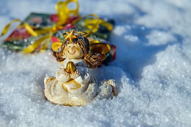 Cheerful angel figurine with golden wings sitting in snow near holiday gift with ribbon. Ideal for use in Christmas greeting cards, festive decoration advertisements, winter holiday promotions, or seasonal social media posts.