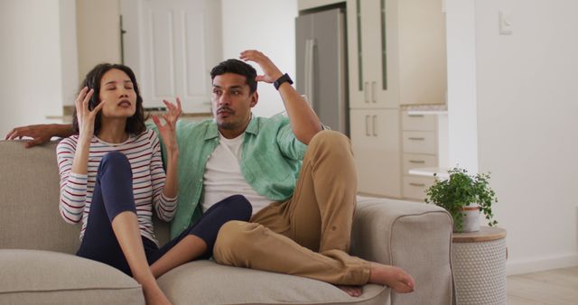 Image shows young couple having a heated discussion while sitting on couch in a modern living room. Ideal for illustrating relationship issues, communication problems, and domestic life themes in articles or blog posts.