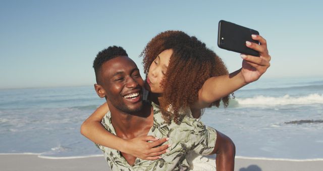 Happy diverse couple piggybacking and taking selfies with smartphone on sunny beach by the sea. Summer, free time, relaxation, communication, romance and vacations.