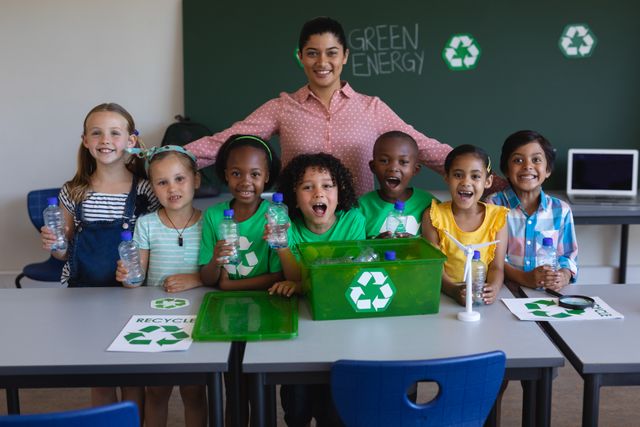 Teacher and diverse group of schoolchildren smiling and holding plastic bottles in classroom. Green energy and recycling symbols on chalkboard and bins. Ideal for educational materials, environmental campaigns, and school programs promoting sustainability.