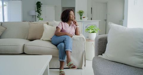 Contemplative woman sitting on beige sofa, gazing left in bright minimalist living room
