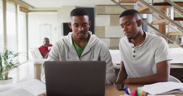 Two african american teenage twin brothers using a laptop and talking with father in background. family leisure time at home together during quarantine lockdown.