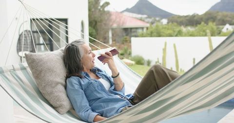 Mature woman relaxing in striped hammock holding smartphone poolside on covered patio