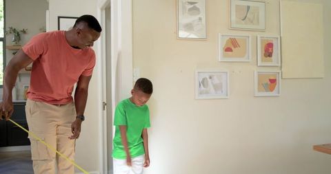 African american father measuring son height with tape measure on home wall, family milestone