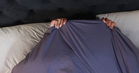 Female hiding under slate purple sheet gripping tufted headboard with white manicured hands