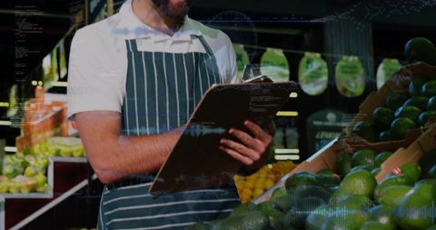 Checking inventory with clipboard in produce aisle, bearded grocery worker noting avocados