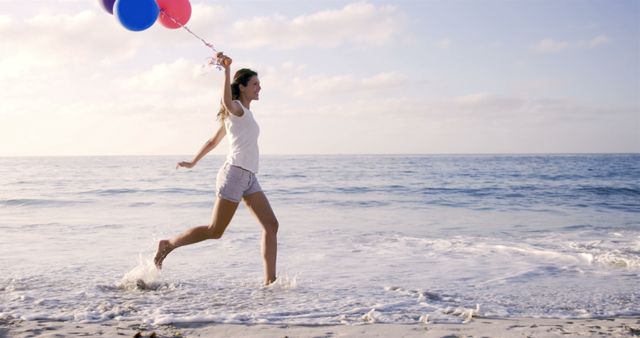 Young woman running along the shoreline while holding colorful balloons, splashing water with feet. Ideal for summer vacation, outdoor recreation, or freedom-themed projects. Perfect for promotions related to travel, leisure, and happiness.