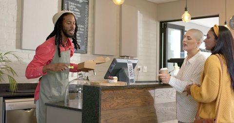 Friendly barista handing takeaway pastry to two customers at modern cafe counter