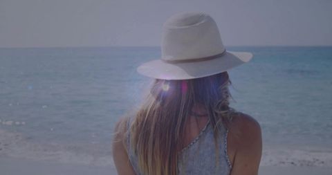 Woman wearing white wide hat gazing at calm ocean horizon with pastel lens flare