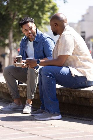 Two men sitting on ledge chatting over coffee in sunny urban scene