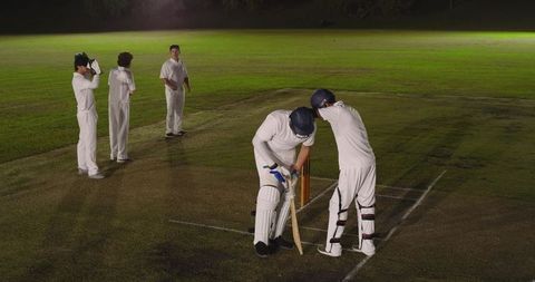 Night cricket match featuring diverse male players, two batsmen at crease under floodlights