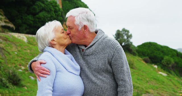 A senior Caucasian couple shares a tender kiss outdoors, with copy space. Their affectionate moment captures a lifetime of love and companionship.