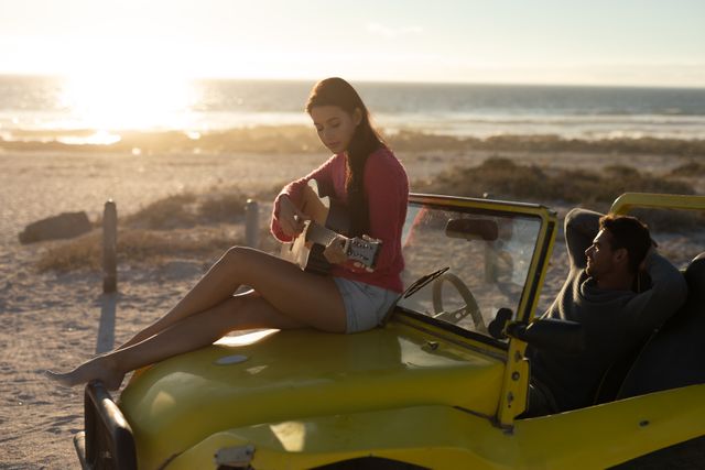 Happy caucasian couple with beach buggy on beach at sunset, woman sitting on car playing guitar. beach stop off on romantic summer holiday road trip.