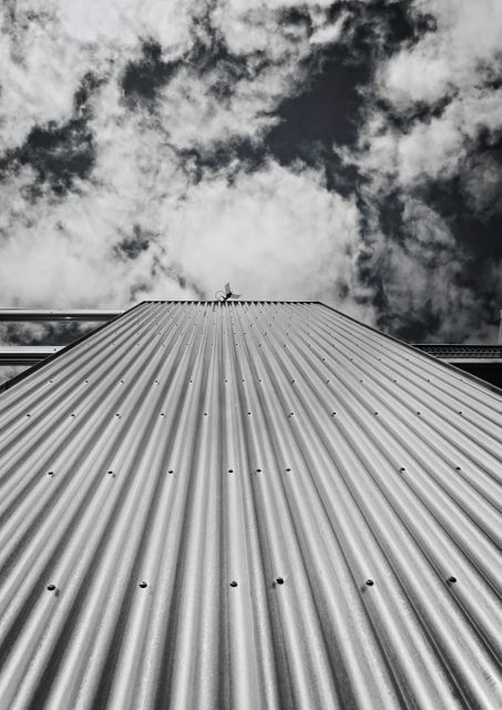 Corrugated metal industrial roof rising towards sky with dramatic clouds forming at the top in black and white. Suitable for use in industrial design, architecture presentations, and as a background for urban-themed projects.