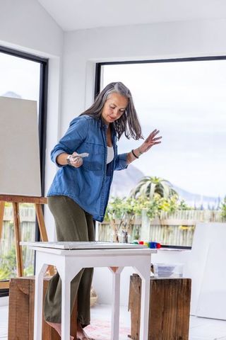 Senior woman artist painting barefoot in bright studio with garden view using palette knife