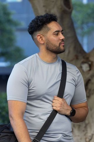 Young man standing in park wearing gray athletic shirt holding crossbody bag with smartwatch
