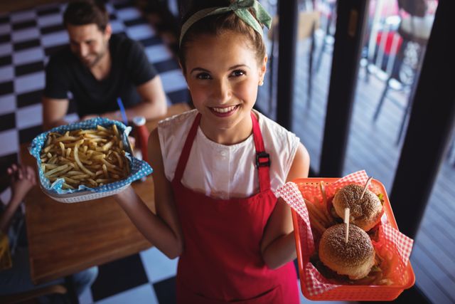 Smiling waitress holding trays with burgers and fries in a casual diner. Ideal for use in articles or advertisements related to the food service industry, customer service, hospitality training, or restaurant promotions.