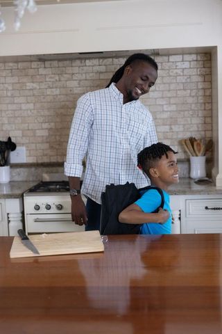 African american father helping son with backpack at kitchen island morning routine