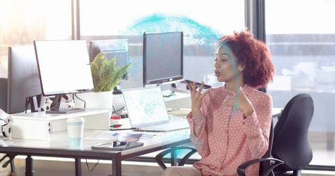 Woman speaking into smartphone while gesturing at monitors in bright open office