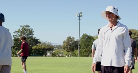 Coach watching team practice on sunny grass field wearing white bucket hat