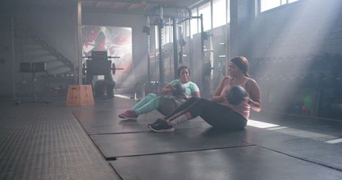 Diverse female friends doing medicine ball core twists in sunlit gym for strength training