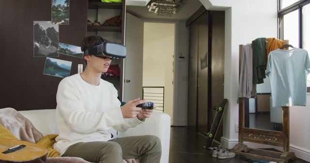 A young man is using a virtual reality headset and a game controller in a modern living room. He is focused on the gaming experience, surrounded by casual items such as a guitar, skateboard, and clothes on a standing mirror. This image can be used to depict the latest in home entertainment technology, immersive experiences, and modern lifestyle trends.