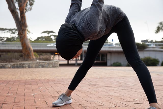 Fit biracial woman wearing hijab and sportswear exercising outdoors in the city, stretching in urban park. Urban lifestyle exercise.