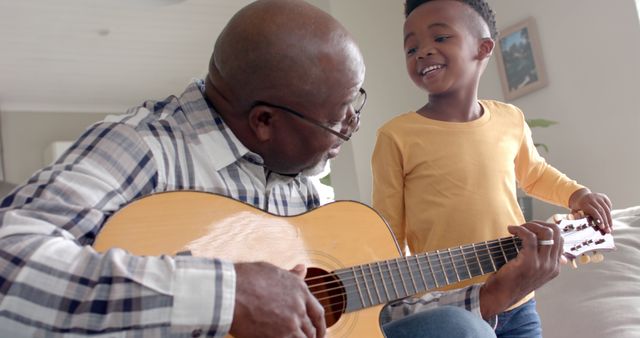 This image captures a heartwarming moment between a grandfather and grandson as the elder teaches the young boy to play guitar at home. Ideal for use in articles or marketing materials focusing on family, music education, elderly role models, and multigenerational activities.