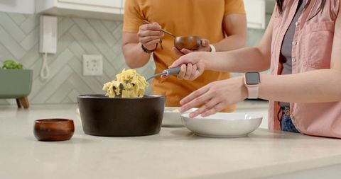 Asian couple serving pasta from black pot on kitchen island with herringbone backsplash