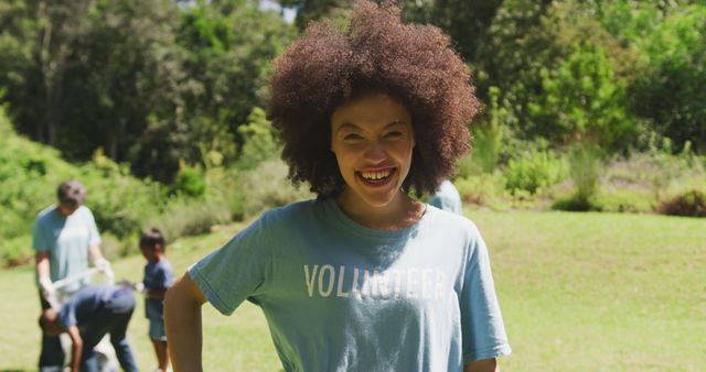 Female volunteer with curly hair smiling during outdoor community service. Others are working in the background. Suitable for projects highlighting volunteerism, community involvement, charity work, and outdoor social events.