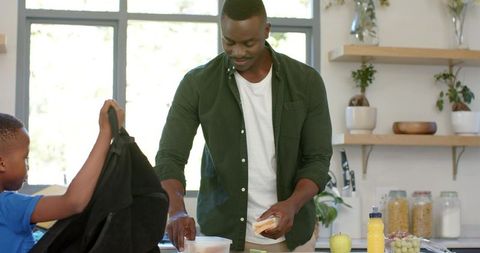 African american father packing healthy school lunch with son in bright home kitchen