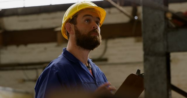 A middle-aged Caucasian man in a yellow hard hat and blue workwear holds a clipboard, with copy space. His focused expression and attire suggest he may be a construction worker or engineer assessing the site.