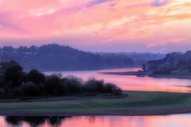 Beautiful illustration of sunrise over a calm river reflects vibrant pink and purple hues in the morning sky. Trees bordering the river and hills in the distance. Perfect for promoting travel destinations, nature blogs, or inspiring relaxation. Ideal for desktop wallpapers, calendar designs, and meditation visuals.