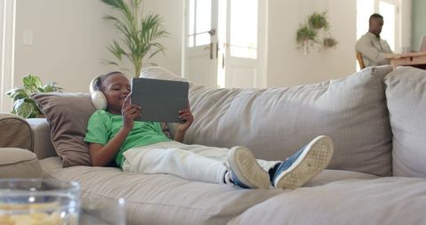 African American Father Working While Son Relaxing on Couch Using Tablet and Headphones