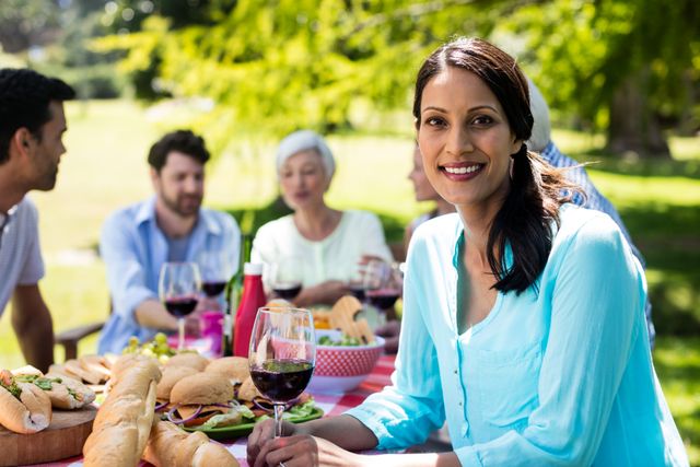 Woman smiling while holding a glass of red wine at a picnic table with friends in a park. Ideal for use in advertisements for outdoor activities, social gatherings, summer events, and lifestyle promotions.