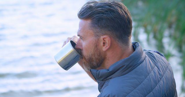 Mature man drinking from a mug while standing by a tranquil lake, wearing a casual jacket. This image is ideal for use in advertisements promoting outdoor activities, relaxation products, travel experiences, or lifestyle content focused on men over 40.