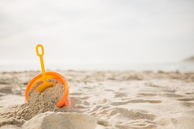 Bright orange bucket filled with sand and a yellow spade on a tropical beach. Ideal for themes related to summer vacations, childhood fun, beach activities, and outdoor leisure. Perfect for travel brochures, family holiday promotions, and children's activity advertisements.